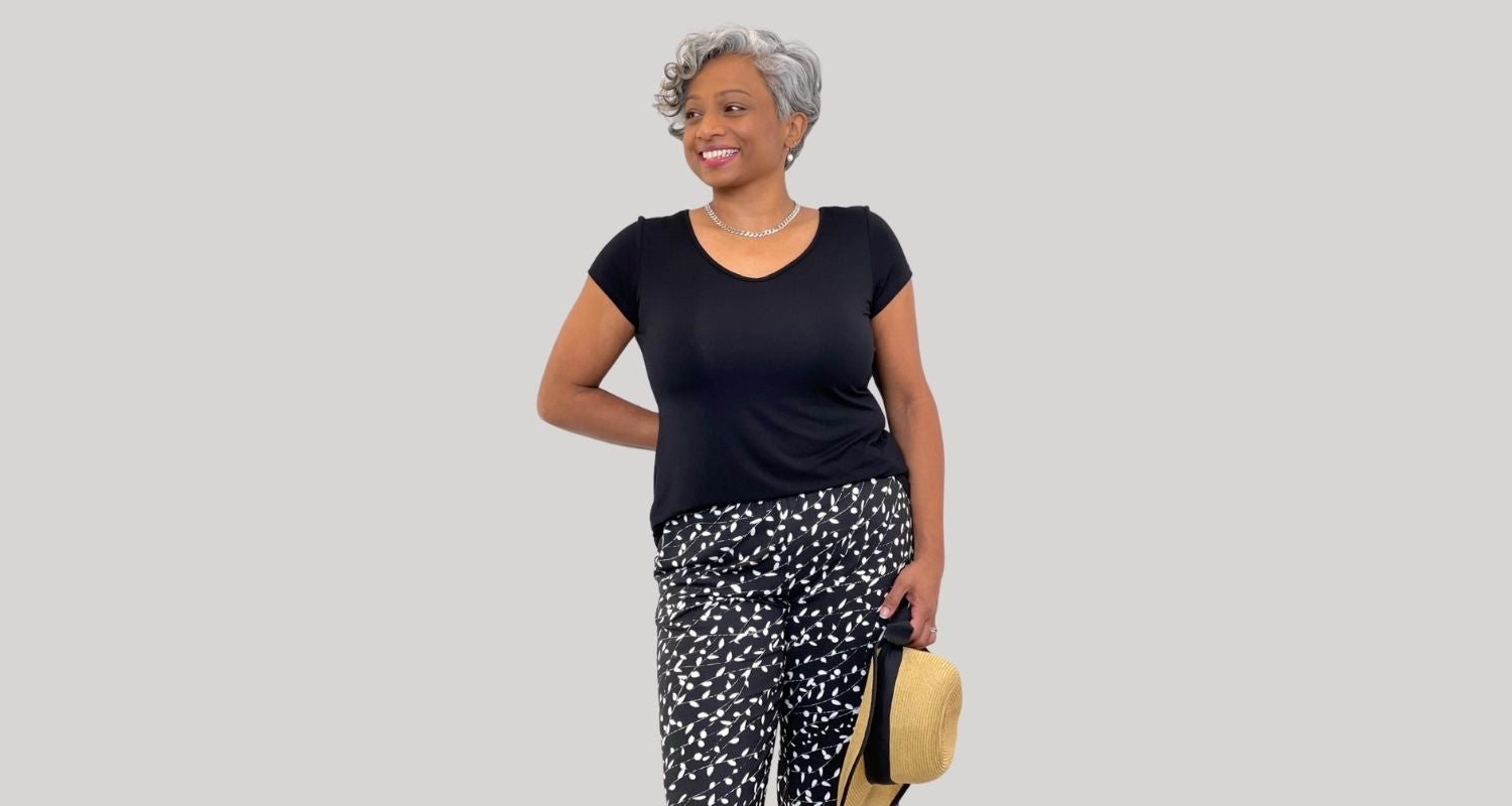 A woman is shown wearing a black Tshirt and some pants with a black and white pattern. She holds a straw hat and looks to the side.