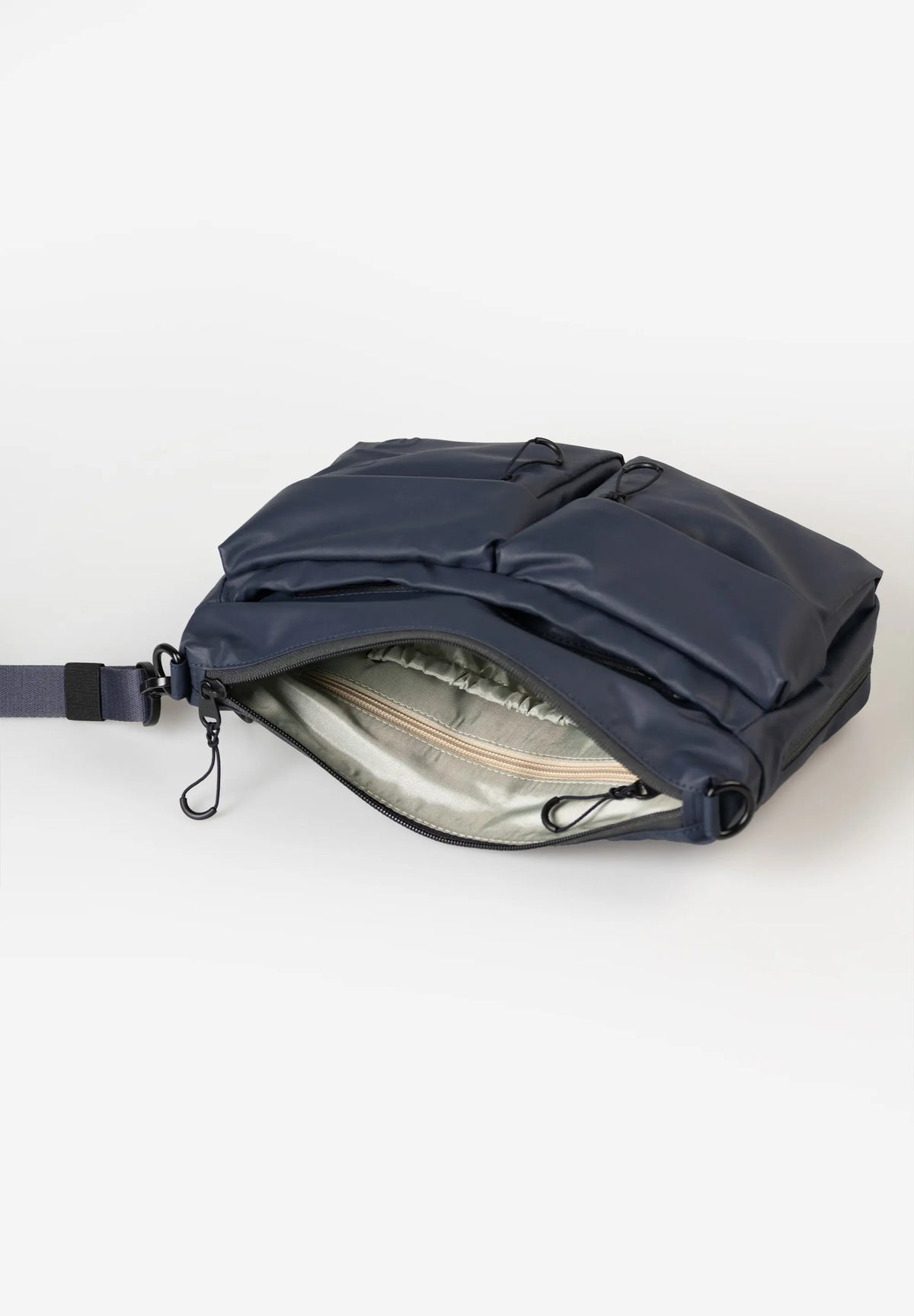 Aunts & Uncles Mogami RE Lite Crossbody Bag. A navy blue crossbody bag with an open front zipper, showing a silver-gray interior lining, photographed on a clean white studio background.