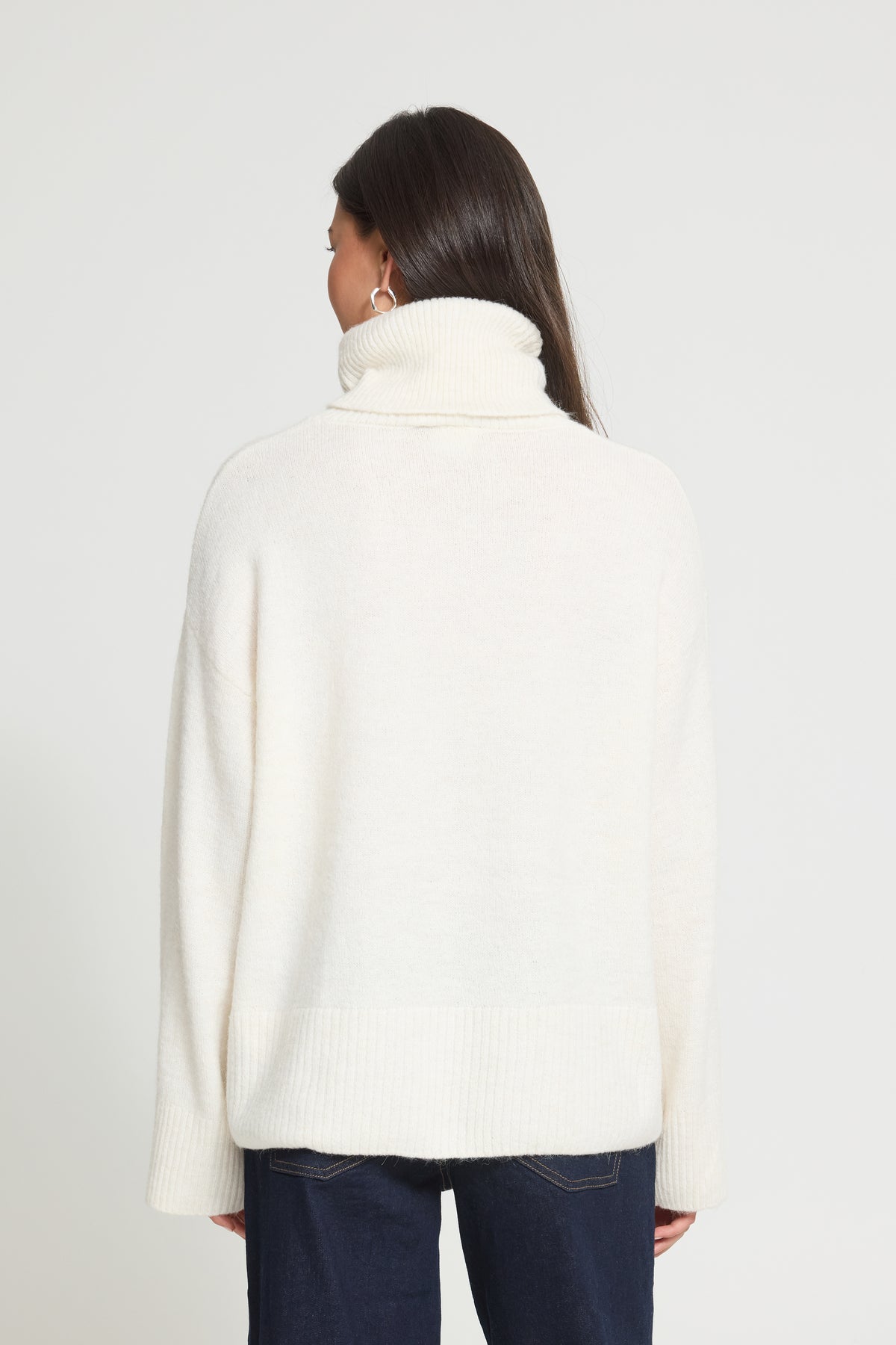 Ichi Kamara Roll Neck Sweater A woman shown from the back wearing a cream-colored, high-neck knit sweater and dark blue jeans against a plain light gray background.