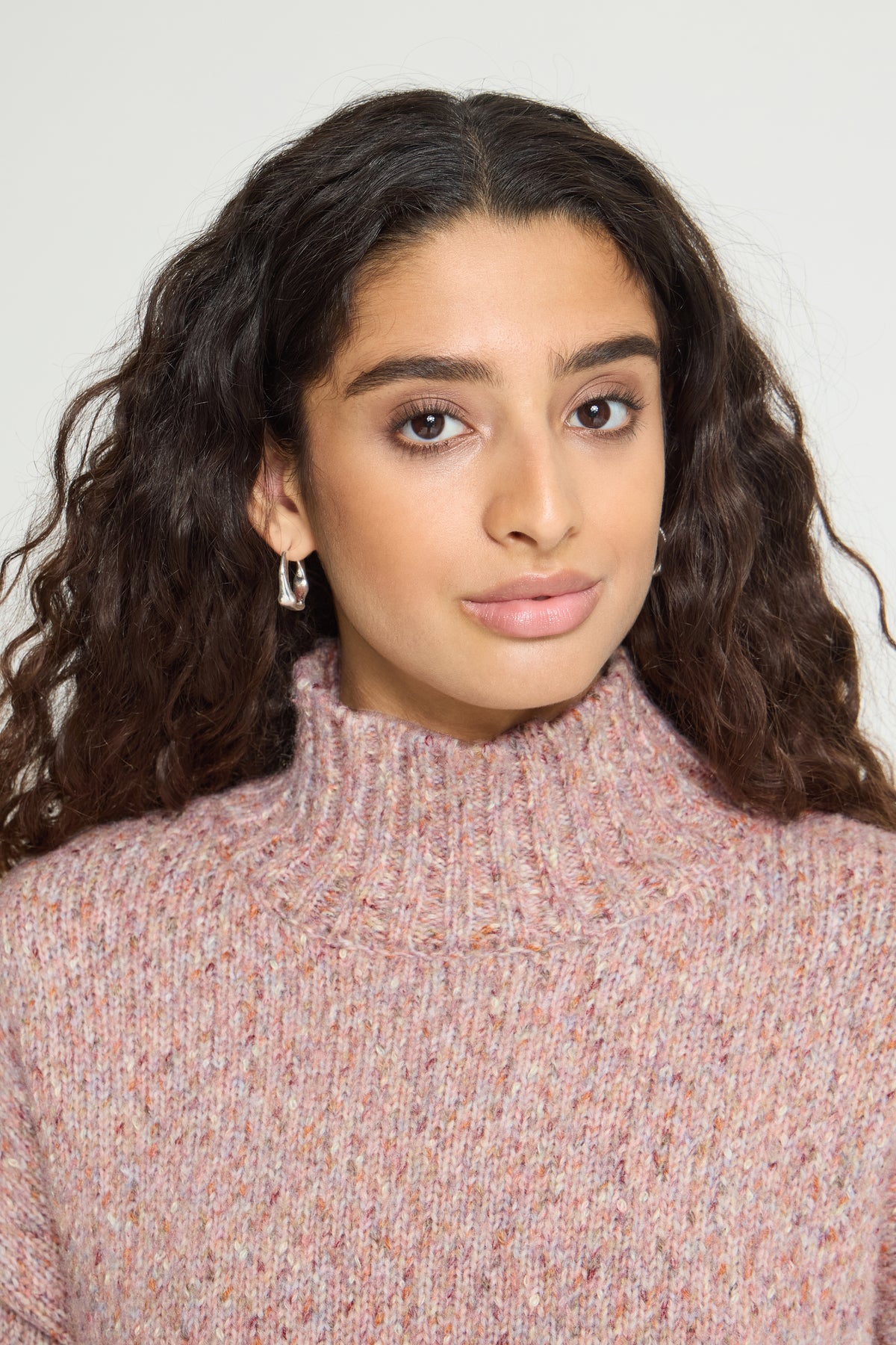 Ichi Fanny Sweater. A close-up portrait of a woman with long, dark, curly hair wearing a textured, heathered pink turtleneck sweater against a plain light background.