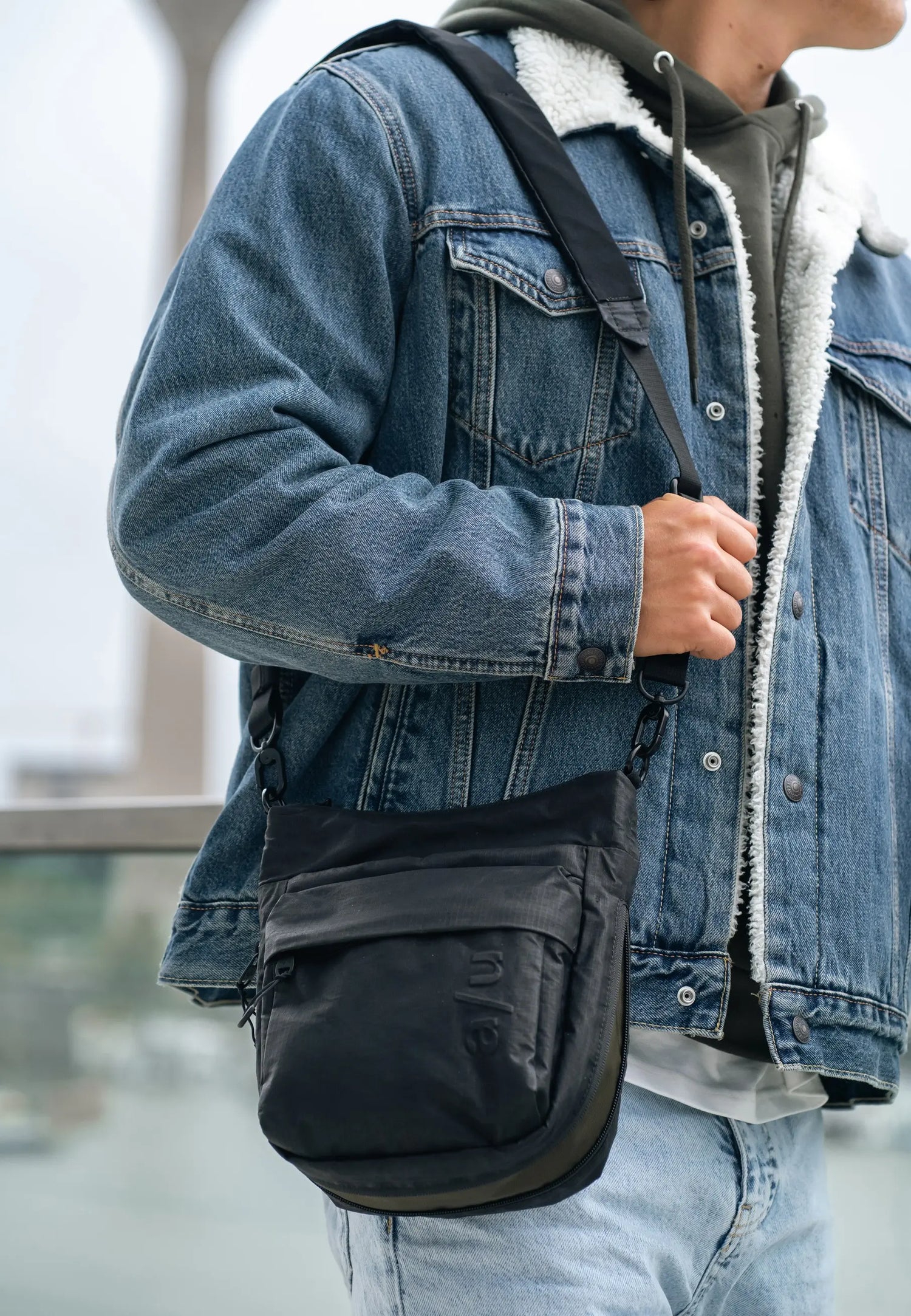 Aunts & Uncles Yura RE Lite Crossbody Bag. A person wearing a blue denim jacket with a white sherpa lining and light blue jeans, carrying a black crossbody sling bag with a black strap.