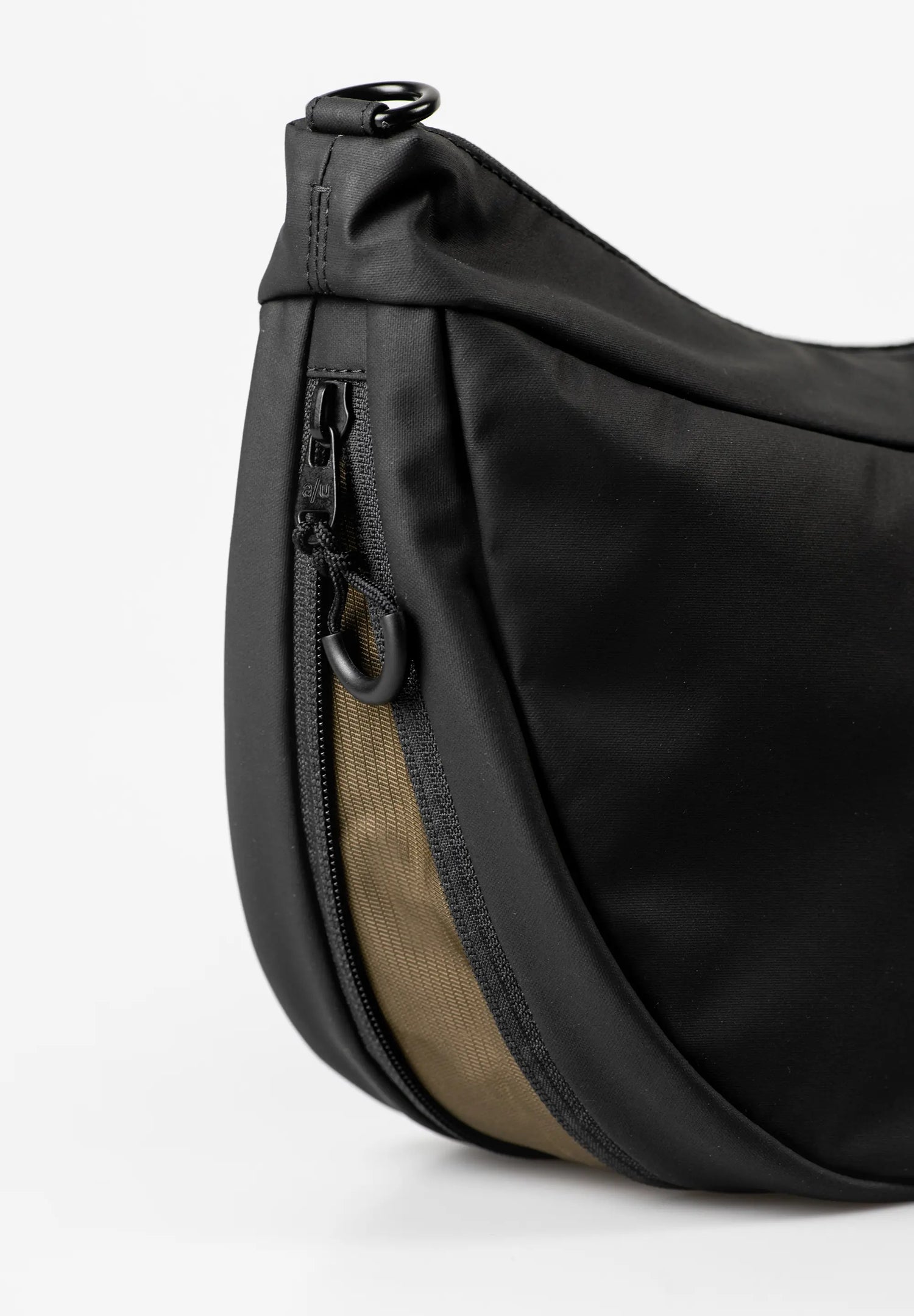 Aunts & Uncles Toyo RE Lite Halfmoon Bag. Close-up of a black nylon waist bag with a tan fabric accent panel and black zipper hardware, photographed on a clean white background.