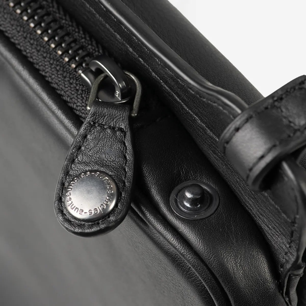 Aunts & Uncles Tulip Leather Phonebag Wallet. Close-up of a black leather bag with a black zipper, metal zipper pull, and a round silver-tone snap button engraved with 'KLEISS-EMPEROR'.