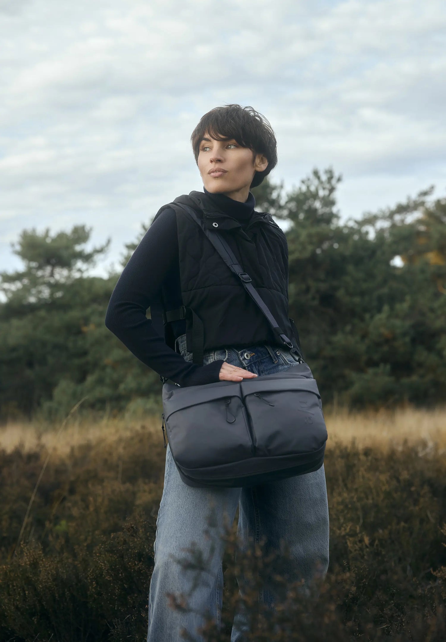 Aunts & Uncles Mogami RE Lite Crossbody Bag. A person standing outdoors in a grassy field under a cloudy sky, wearing a dark gray crossbody messenger bag across their torso.