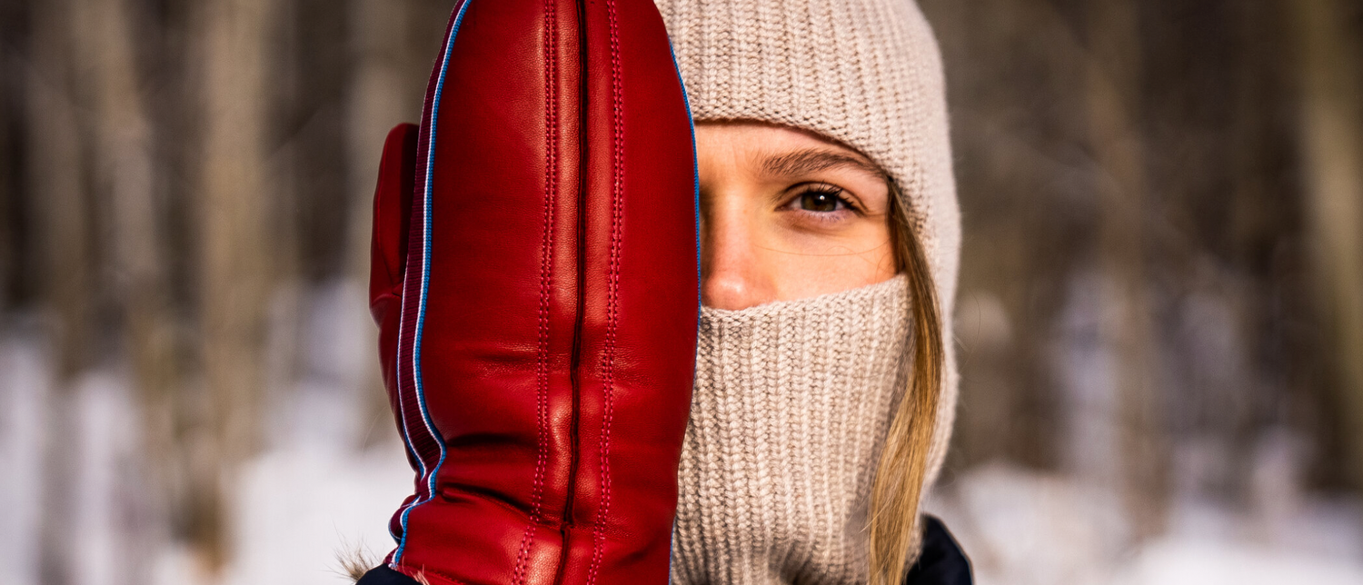 A woman wears a ribbed hat and neckwarmer. She holds a red leather mitt in front of one side of her face.