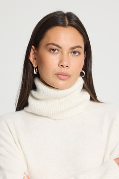 Ichi Kamara Roll Neck Sweater A woman with long dark hair wearing a cream ribbed turtleneck sweater and silver hoop earrings, photographed against a light gray studio background.