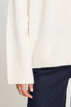 Ichi Kamara Roll Neck Sweater Close-up of a person wearing a cream-colored knit sweater with ribbed cuffs and hem, paired with dark navy pants. The person's hand is visible wearing a silver ring.