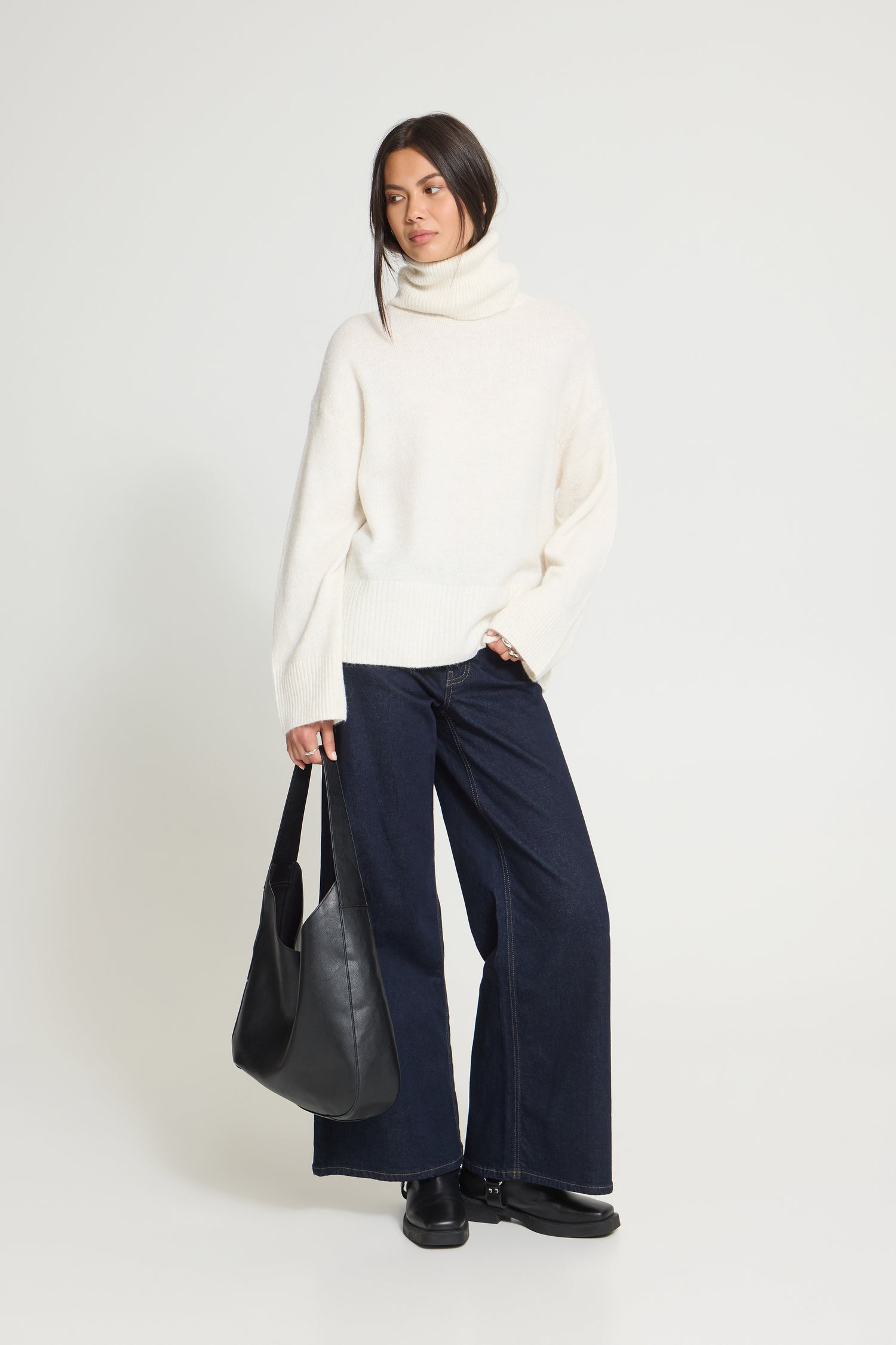 Ichi Kamara Roll Neck Sweater A woman stands against a light gray studio background wearing a cream turtleneck sweater, dark navy wide-leg pants, black shoes, and holding a black leather tote bag.