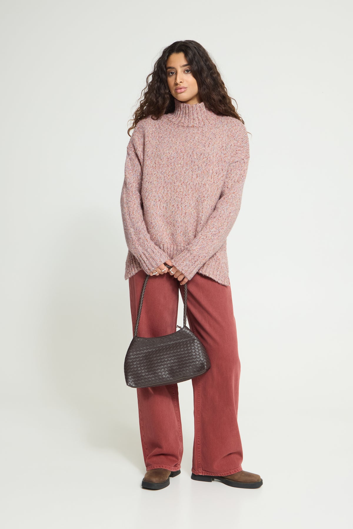 Ichi Fanny Sweater. A woman modeling a chunky knit turtleneck sweater in dusty rose pink, paired with rust-colored wide-leg trousers and a dark gray textured shoulder bag, against a plain white studio background.