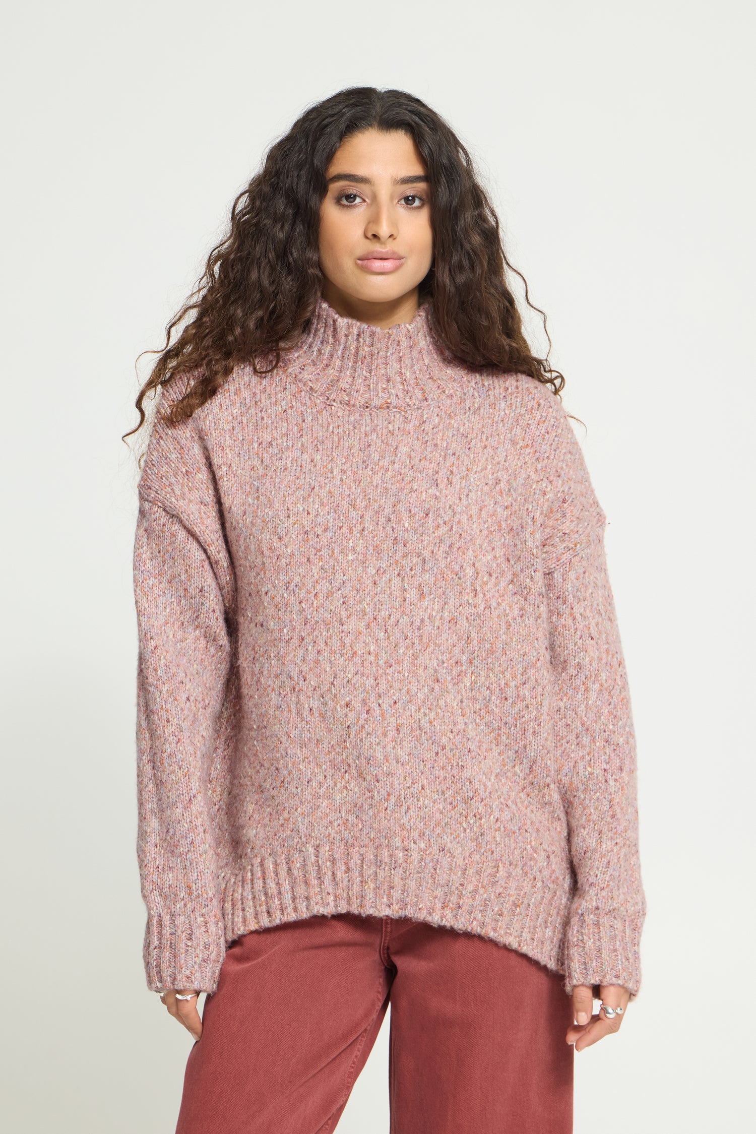 Ichi Fanny Sweater. A woman wearing a chunky, oversized turtleneck sweater in a dusty rose pink color, paired with rust-colored pants, against a plain light gray studio background.