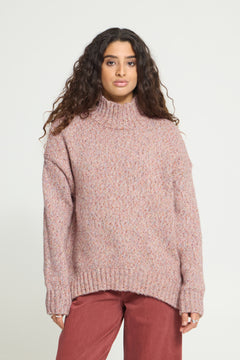 Ichi Fanny Sweater. A woman wearing a chunky, oversized turtleneck sweater in a dusty rose pink color, paired with rust-colored pants, against a plain light gray studio background.