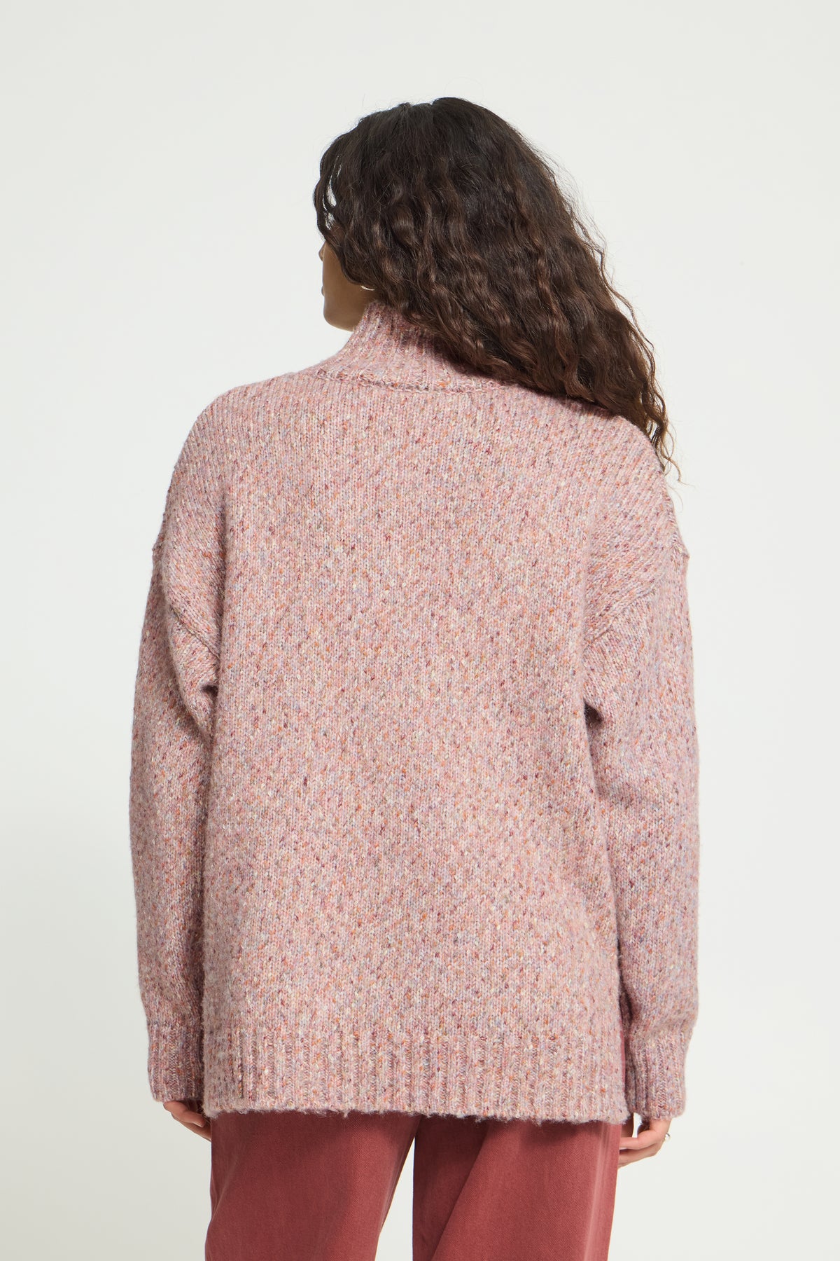 Ichi Fanny Sweater. A woman viewed from the back wearing a chunky knit turtleneck sweater in a dusty rose pink color, paired with rust-colored pants, against a plain white studio background.