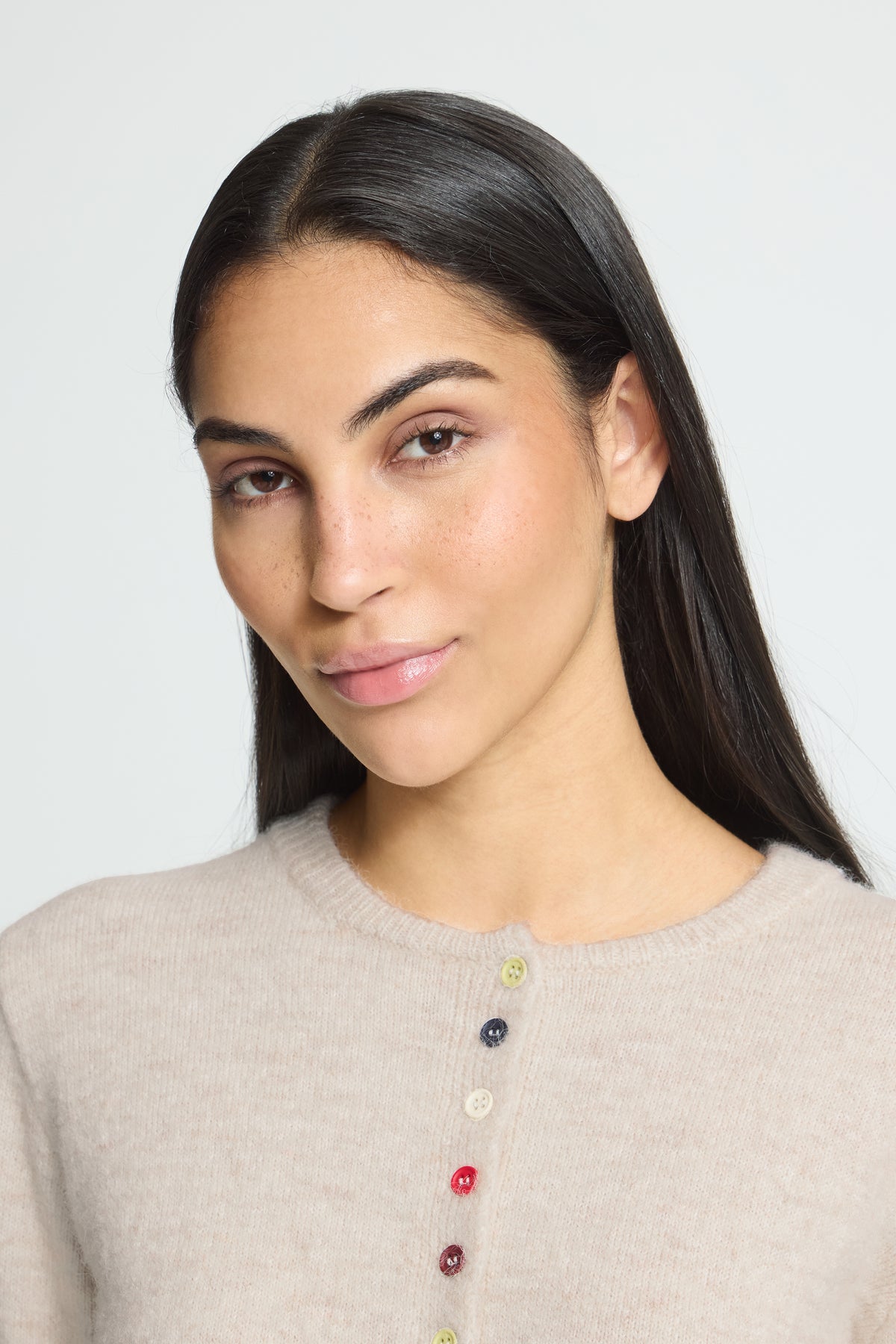 Ichi Fanita Cardigan. A close-up portrait of a woman with long dark hair wearing a light beige knit sweater with colorful decorative buttons down the front, set against a plain light gray background.