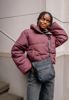 Aunts & Uncles Yura RE Lite Crossbody Bag. A woman wearing a cropped, oversized plum-colored puffer jacket and blue jeans, carrying a dark slate blue crossbody bag. She is posing on outdoor concrete stairs against a light gray tiled wall.