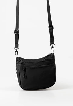 Aunts & Uncles Nanuki RE Lite Crossbody Bag. A black nylon shoulder bag with an adjustable, detachable black strap, featuring a front zippered pocket, photographed against a clean white studio background.