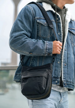 Aunts & Uncles Yura RE Lite Crossbody Bag. A person wearing a blue denim jacket with a white sherpa lining and light blue jeans, carrying a black crossbody sling bag with a black strap.