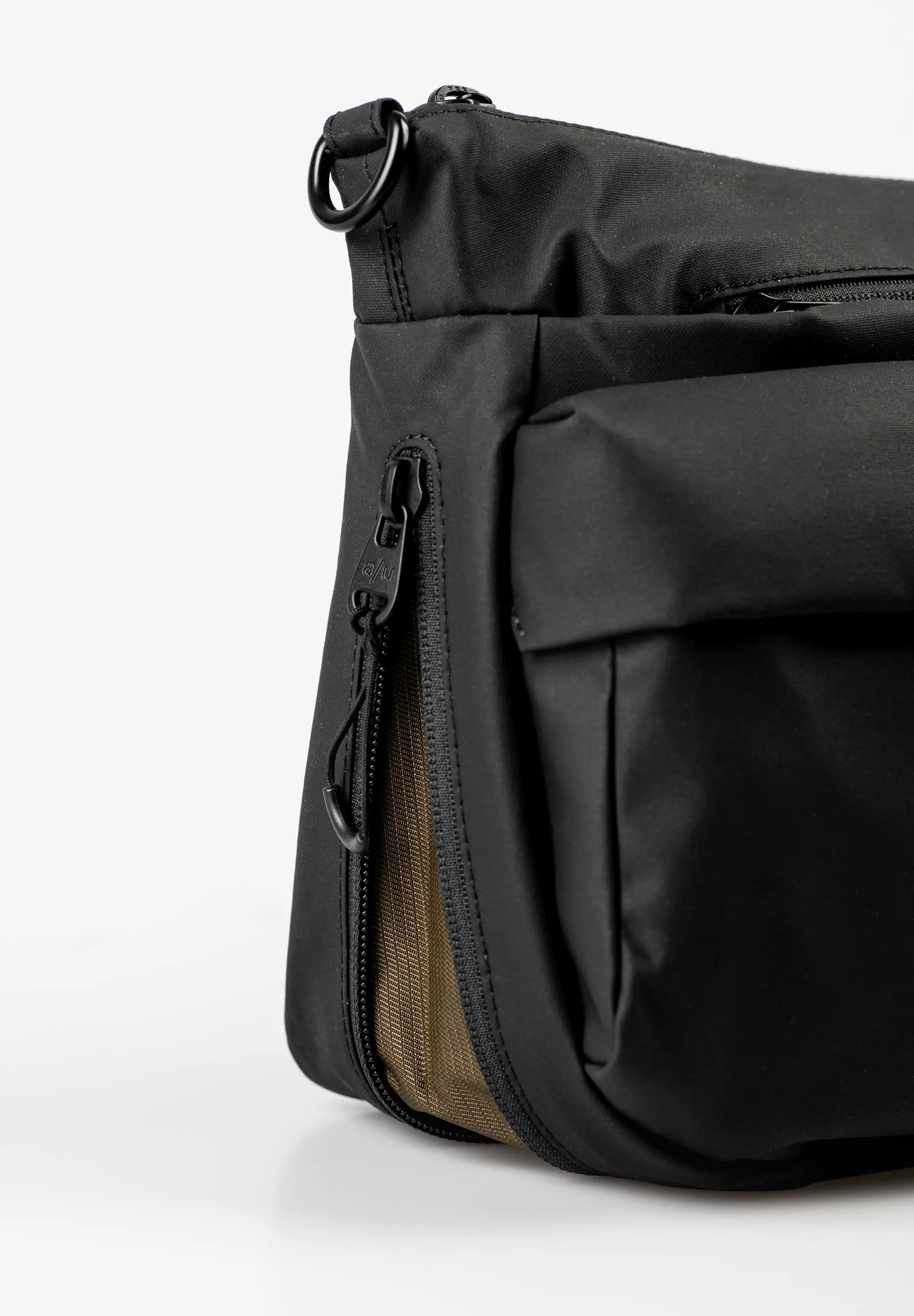 Aunts & Uncles Mogami RE Lite Crossbody Bag. Close-up of a black nylon shoulder bag showing a side zippered pocket with a tan mesh lining and a black metal D-ring attachment.