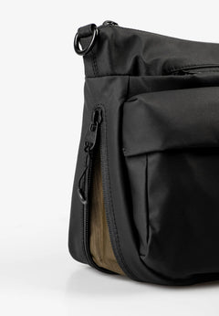 Aunts & Uncles Mogami RE Lite Crossbody Bag. Close-up of a black nylon shoulder bag showing a side zippered pocket with a tan mesh lining and a black metal D-ring attachment.