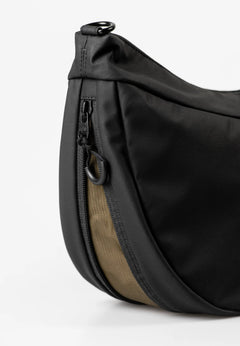Aunts & Uncles Toyo RE Lite Halfmoon Bag. Close-up of a black nylon waist bag with a tan fabric accent panel and black zipper hardware, photographed on a clean white background.