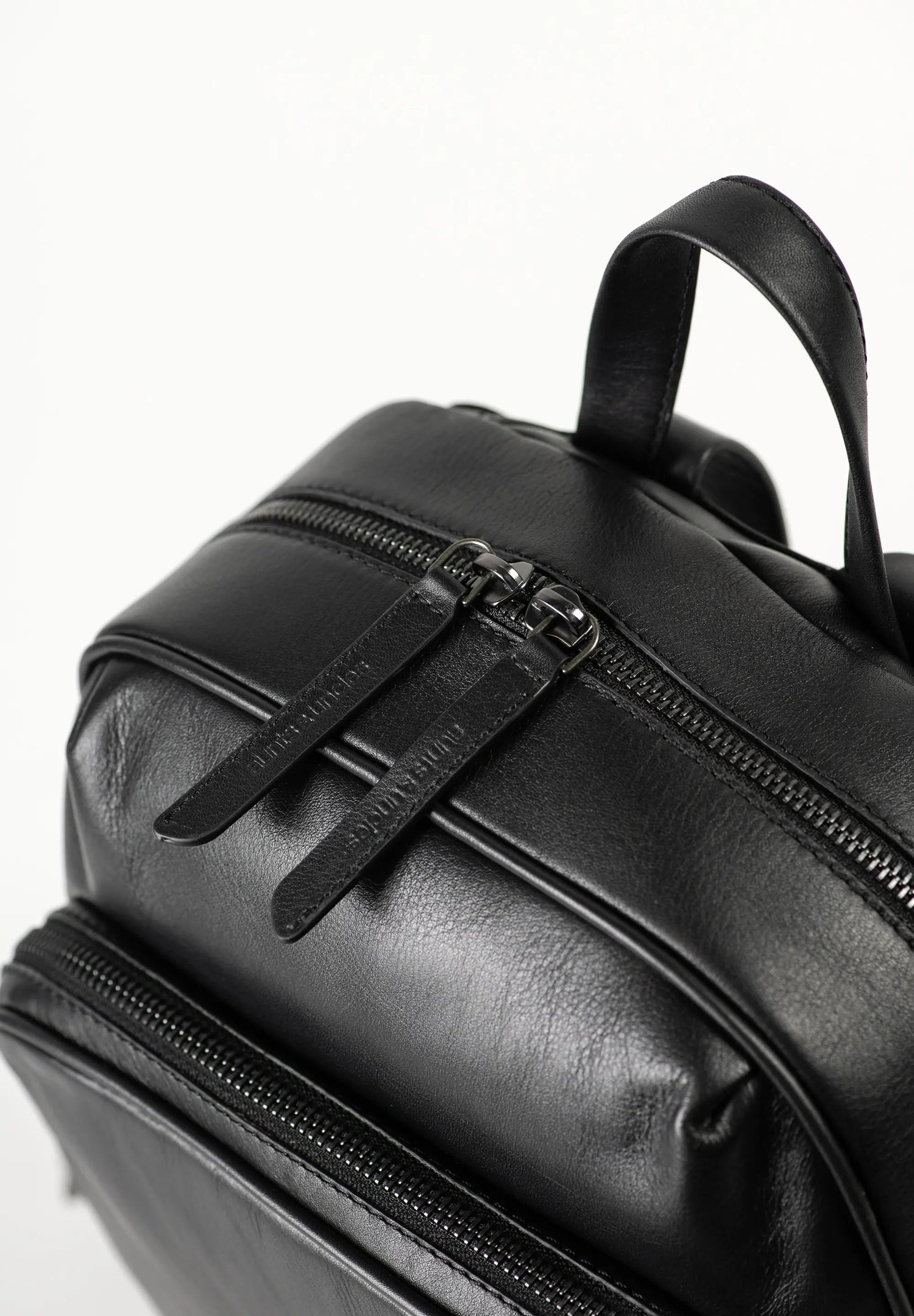Aunts & Uncles Coco Leather Backpack Purse.  Close-up of a black leather backpack showing a front zippered pocket with a pull tab and a top carry handle against a white background.