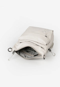 Aunts & Uncles Yura RE Lite Crossbody Bag. A light beige puffer-style pouch bag shown open, revealing a gray interior lining and silver metal zipper hardware, on a clean white background.