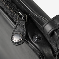 Aunts & Uncles Tulip Leather Phonebag Wallet. Close-up of a black leather bag with a black zipper, metal zipper pull, and a round silver-tone snap button engraved with 'KLEISS-EMPEROR'.