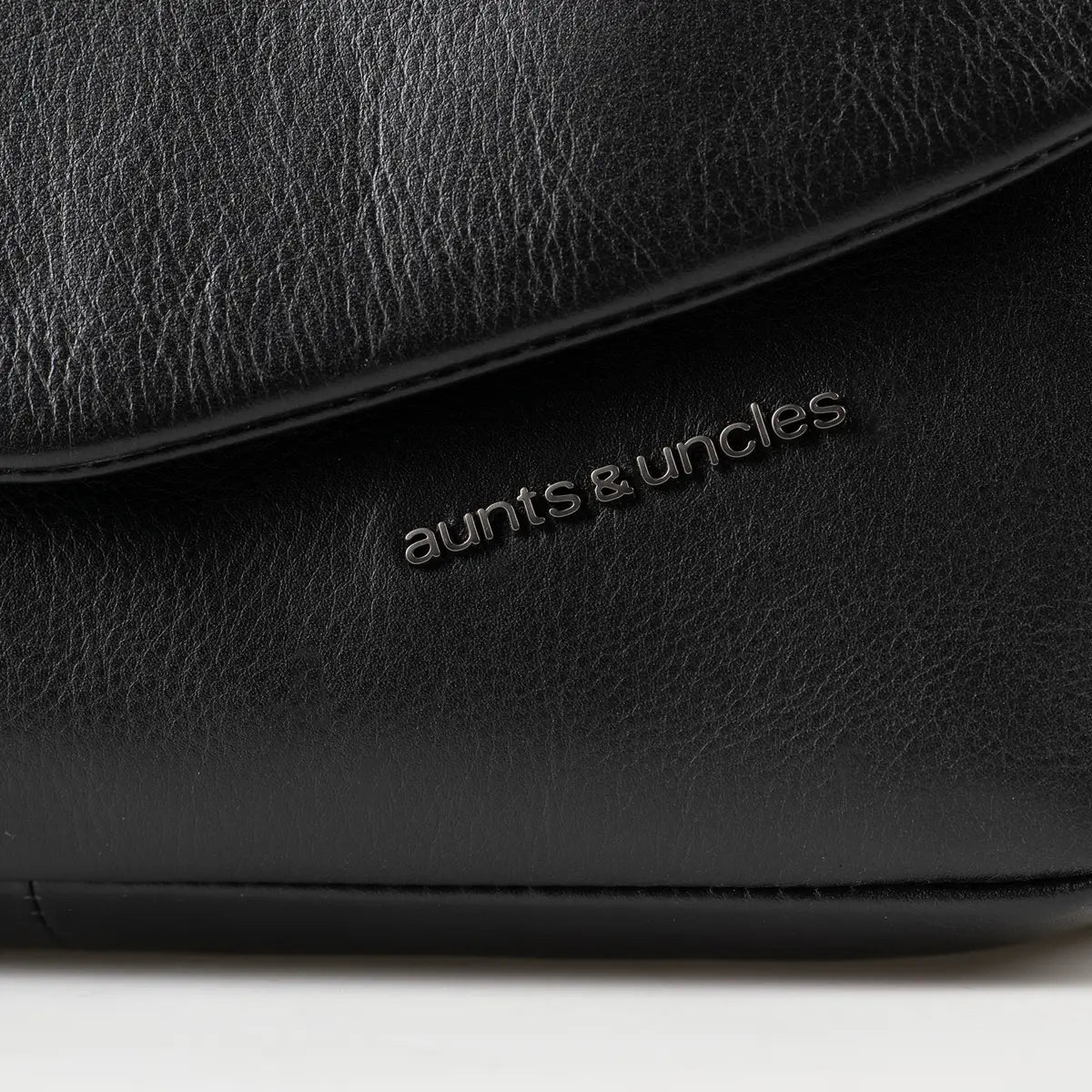 Aunts & Uncles Fern Leather Crossbody Purse. Close-up of a black pebbled leather bag with the 'aunts & uncles' logo debossed on the front flap.