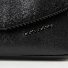 Aunts & Uncles Fern Leather Crossbody Purse. Close-up of a black pebbled leather bag with the 'aunts & uncles' logo debossed on the front flap.
