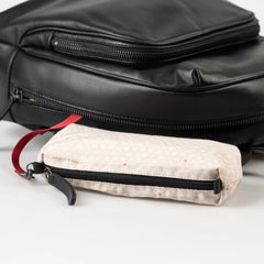 Aunts & Uncles Coco Leather Backpack Purse.  A close-up of a small beige quilted zippered pouch with a subtle pattern, black zipper, and red strap, shown in front of a black bag on a white surface.