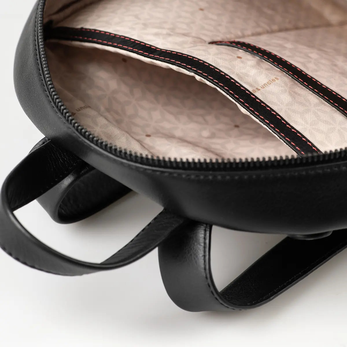 Aunts & Uncles Coco Leather Backpack Purse.  Close-up of the interior of a black leather backpack, showing a beige patterned lining with black and red contrast stitching and zipper details.