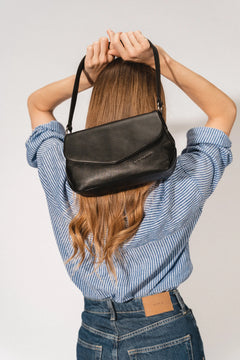 Aunts & Uncles Fern Leather Crossbody Purse. A woman viewed from behind wearing a blue and white striped shirt and jeans, holding a black leather shoulder bag over her head against a light gray background.