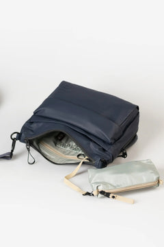 Aunts & Uncles Nanuki RE Lite Crossbody Bag. A navy blue backpack shown open on a light gray studio background, with a small light sage green pouch placed in front of it.