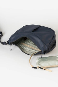 Aunts & Uncles Toyo RE Lite Halfmoon Bag. A navy blue crossbody bag with its front zippered pocket open, showing a light sage green interior lining. A matching sage green zippered pouch is placed beside it on a clean white background.