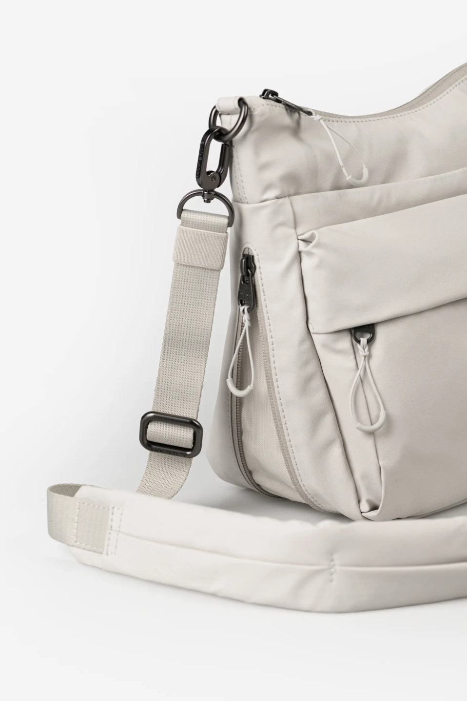 Aunts & Uncles Yura RE Lite Crossbody Bag.A light beige nylon crossbody bag with multiple zippered pockets and an adjustable shoulder strap, shown on a clean white background.