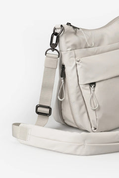 Aunts & Uncles Yura RE Lite Crossbody Bag.A light beige nylon crossbody bag with multiple zippered pockets and an adjustable shoulder strap, shown on a clean white background.