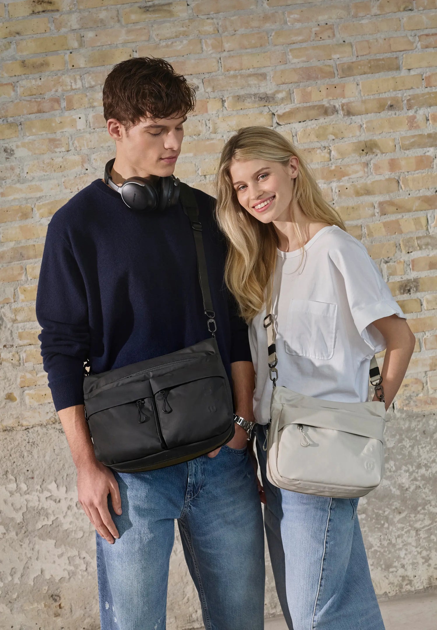 Aunts & Uncles Nanuki RE Lite Crossbody Bag. A man and a woman standing side-by-side against a rustic brick wall, each wearing a messenger bag. The man on the left wears a dark navy sweater and blue jeans, carrying a black leather messenger bag.