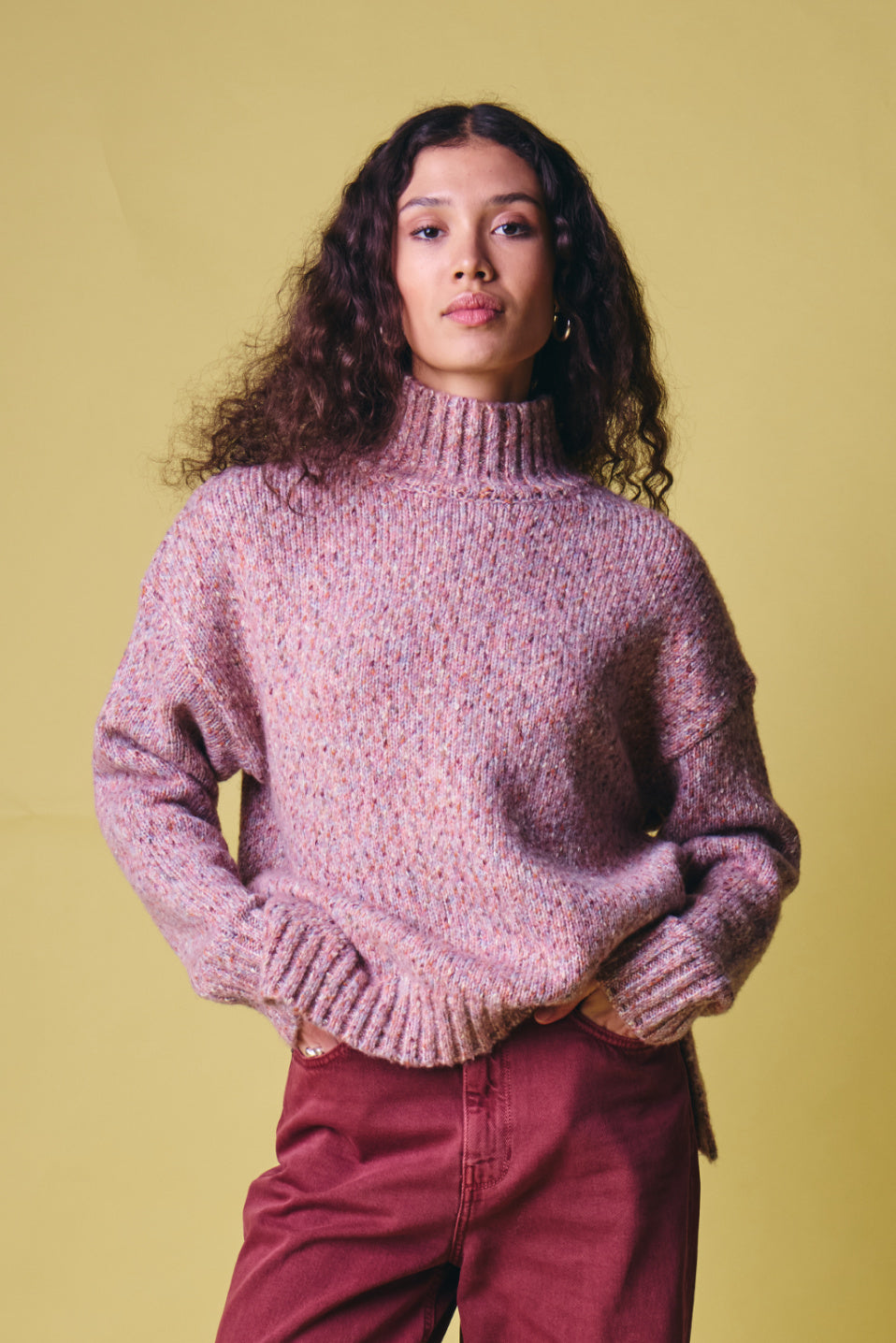 Ichi Fanny Sweater. A woman with long curly dark hair wearing a chunky knit turtleneck sweater in a heathered dusty pink and purple colorway, paired with burgundy pants, standing against a solid pale yellow background.