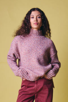 Ichi Fanny Sweater. A woman with long curly dark hair wearing a chunky knit turtleneck sweater in a heathered dusty pink and purple colorway, paired with burgundy pants, standing against a solid pale yellow background.