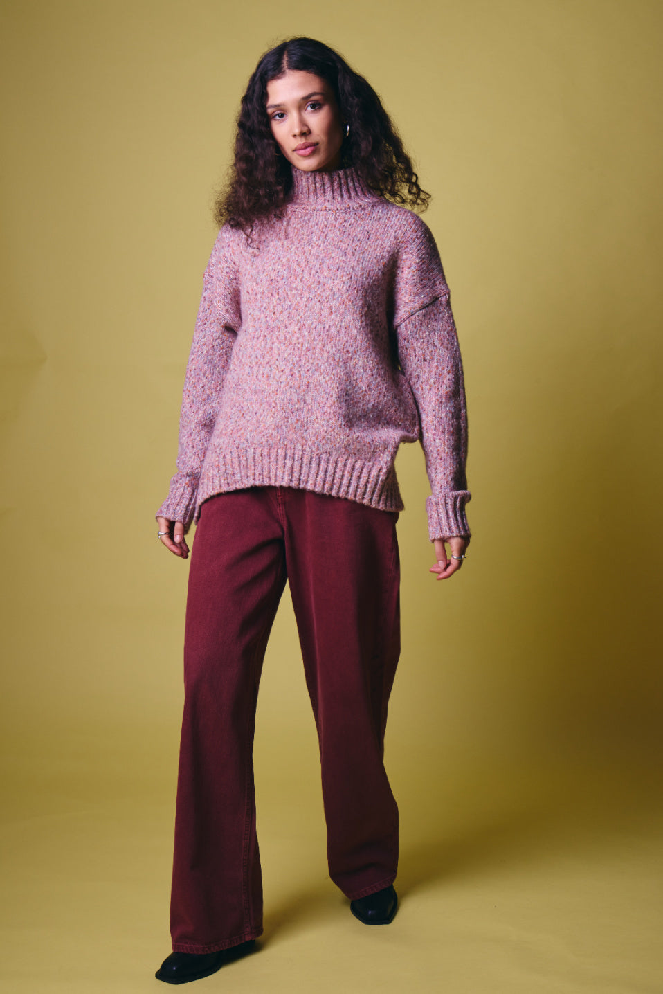 Ichi Fanny Sweater. A model stands against a solid mustard yellow studio background wearing a pink and purple marled knit turtleneck sweater and dark burgundy wide-leg trousers.