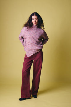 Ichi Fanny Sweater. A model stands against a solid mustard yellow studio background wearing a chunky, textured pinkish-purple turtleneck sweater and wide-leg burgundy trousers.
