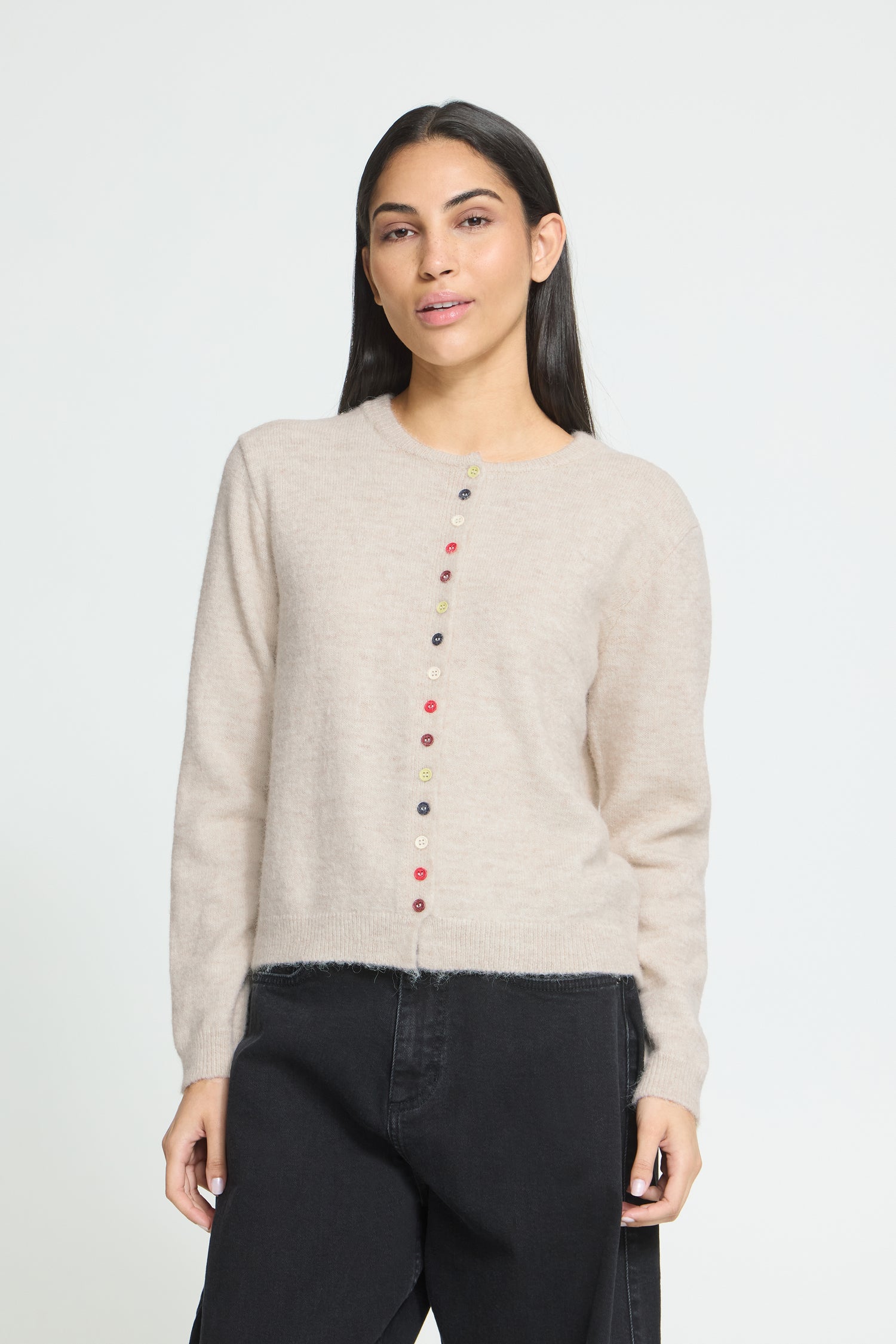 Ichi Fanita Cardigan. A woman modeling a light beige knit sweater with a vertical row of colorful buttons down the front, paired with black pants, against a plain light gray background.