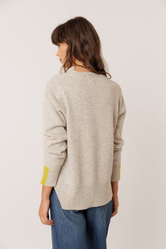 A woman shown from the back wearing a light beige knit sweater with bright yellow accent panels on the cuffs, paired with blue jeans, against a plain off-white background.
