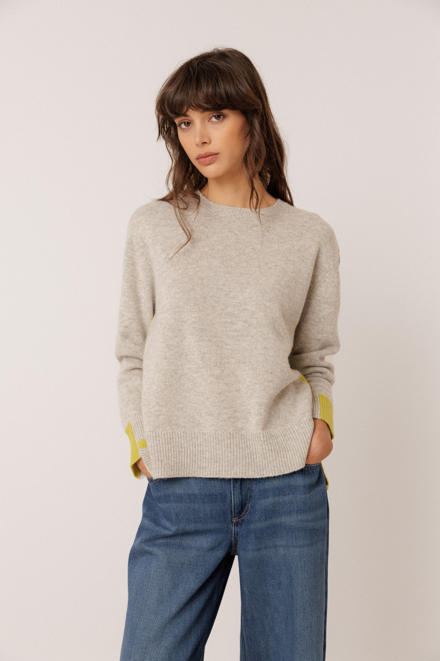 A woman wearing a light beige knit sweater with yellow-green contrast panels on the cuffs, paired with blue jeans, standing against a plain light gray studio background.