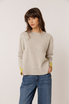 A woman wearing a light beige knit sweater with yellow-green contrast panels on the cuffs, paired with blue jeans, standing against a plain light gray studio background.
