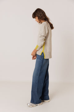 A woman wearing a light beige knit sweater with bright yellow contrast panels on the cuffs and side, paired with wide-leg blue jeans and silver sneakers, standing in profile against a plain off-white studio background.
